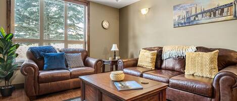 A living room with two brown leather sofas, a wooden coffee table, and a large window with a view of trees.