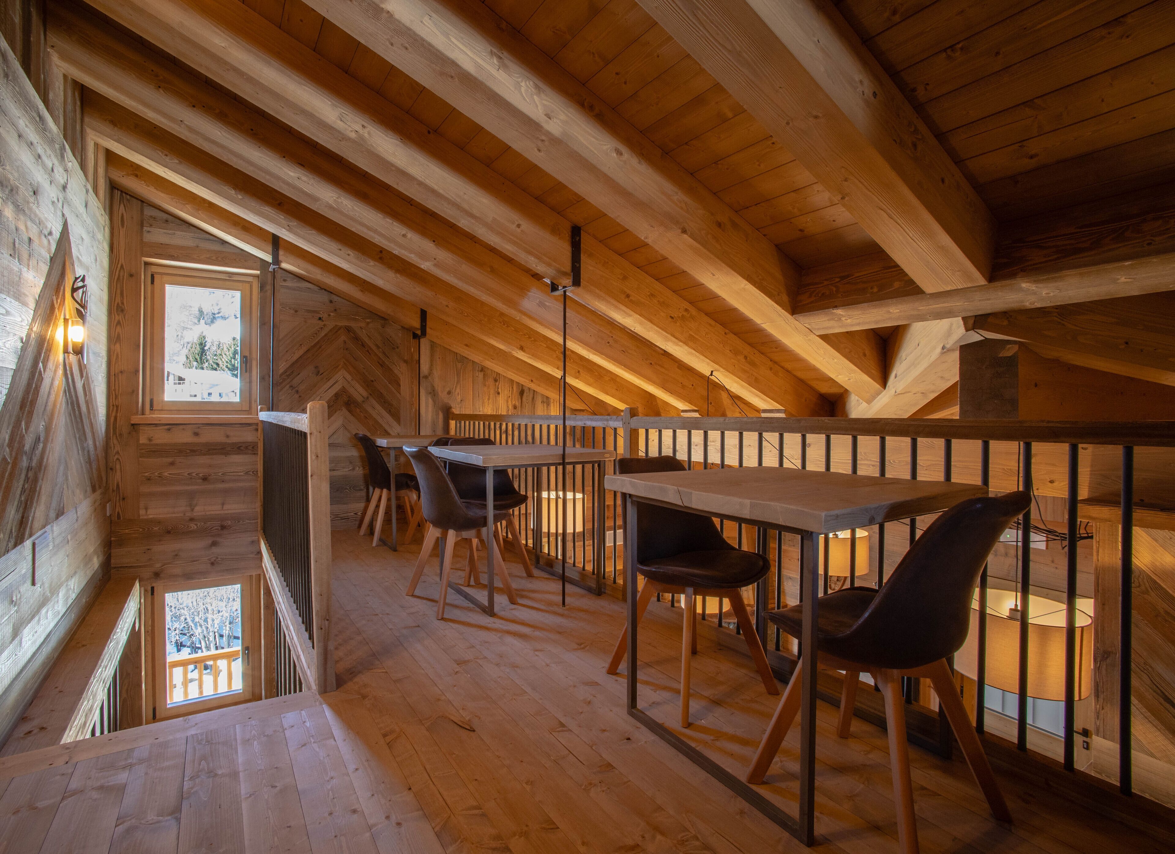 Un interior de madera con un comedor que incluye mesas y sillas, una barandilla, y vistas a un paisaje nevado a través de una ventana.