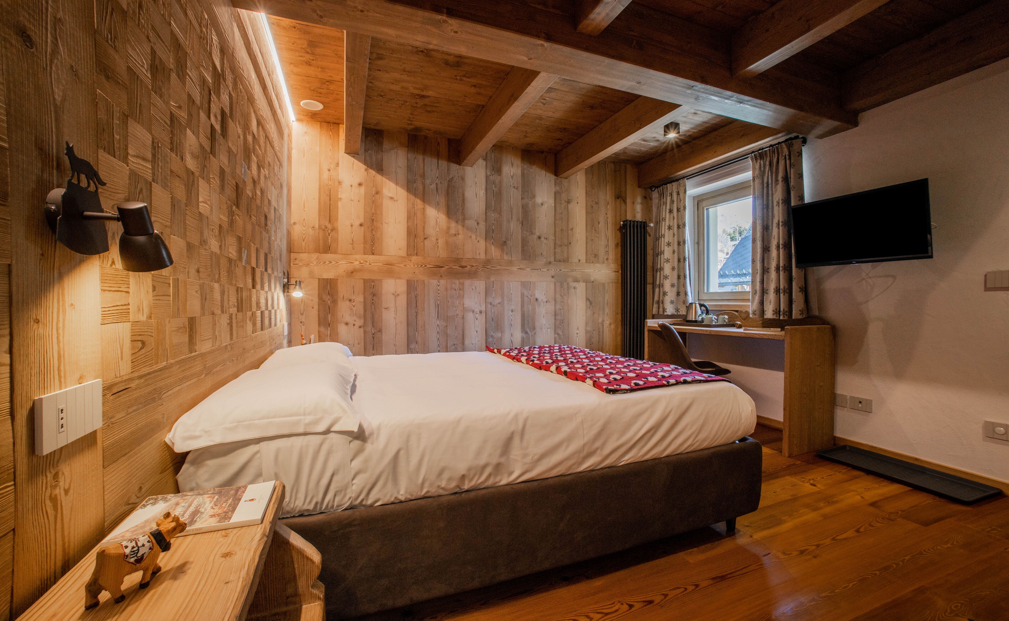 Ein Schlafzimmer mit Holzbalkendecke, einem Bett mit weißer Bettwäsche, einem Schreibtisch mit Computer und einem an der Wand befestigten Fernseher.