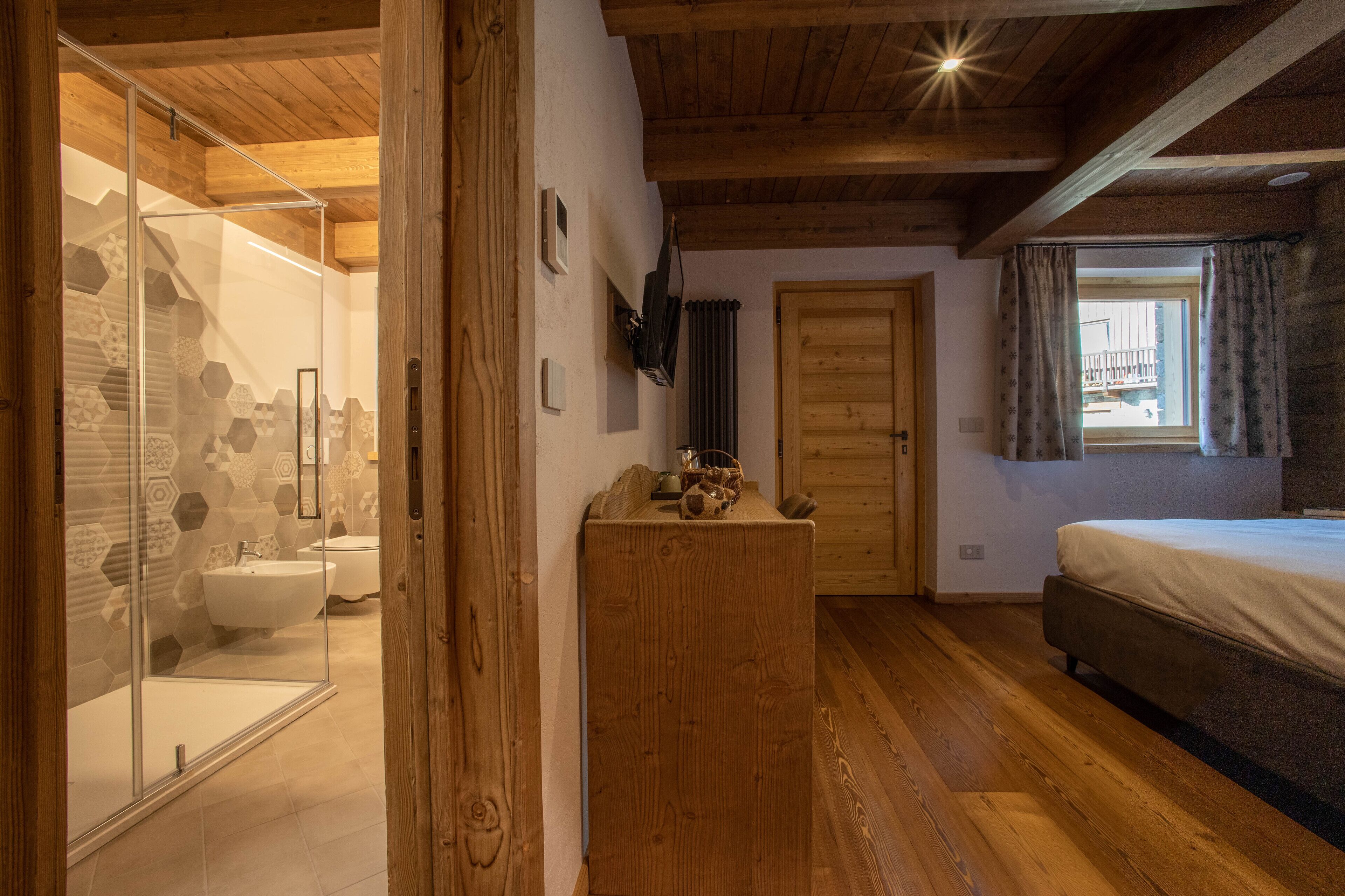 A bedroom with a wooden ceiling, a bed, a wooden dresser, and a bathroom with a glass shower enclosure and a hexagonal tile wall.