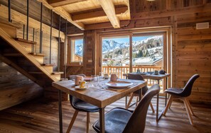 A wooden cabin interior with a dining table set for two, a staircase, and a view of snowy mountains through large windows. - Fleur des Neiges (Ayas)