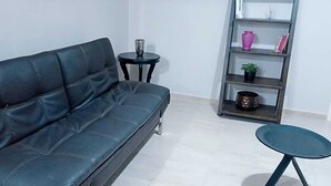 A modern living room with a black leather sofa, a small round table, a tall bookshelf, and a potted plant. - Exclusive apartment in Mxl 3 (Mexicali)