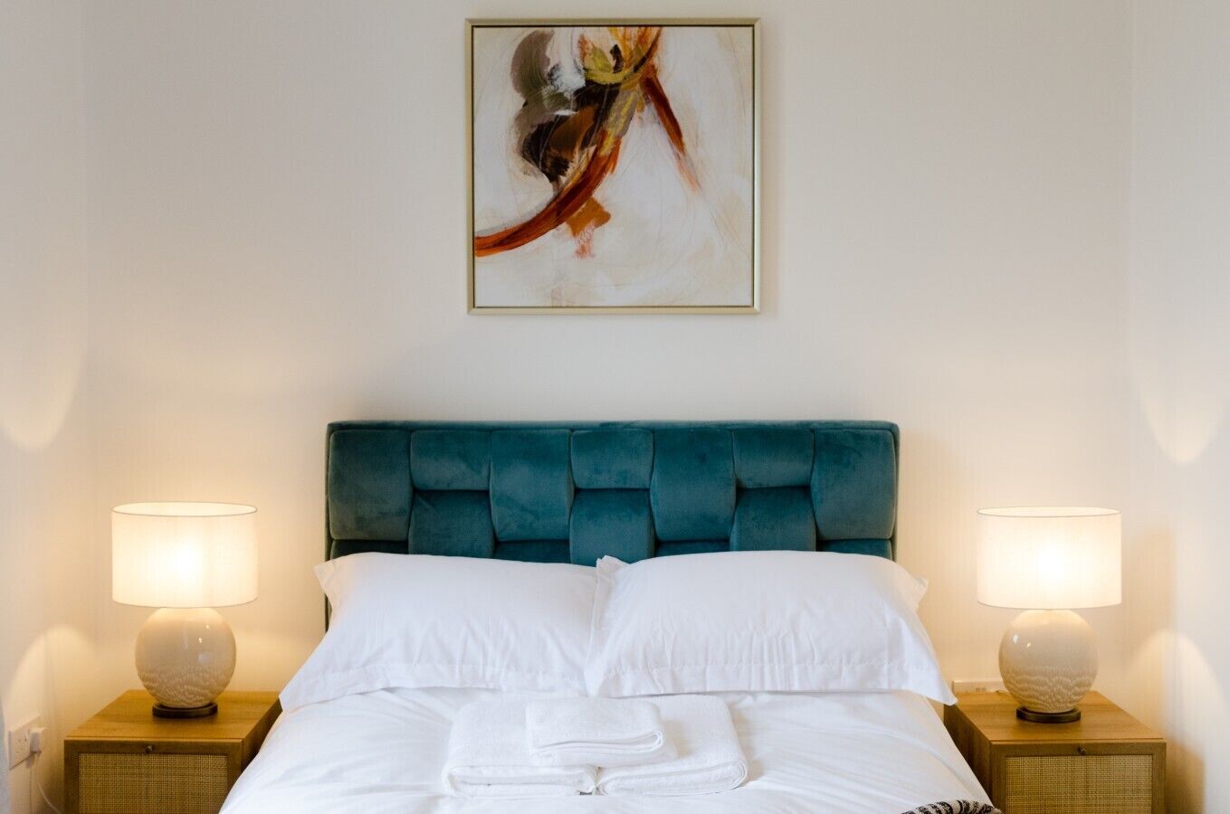 A hotel room with a large bed, two bedside lamps, a wall-mounted abstract painting, and a neatly made bed with folded towels.