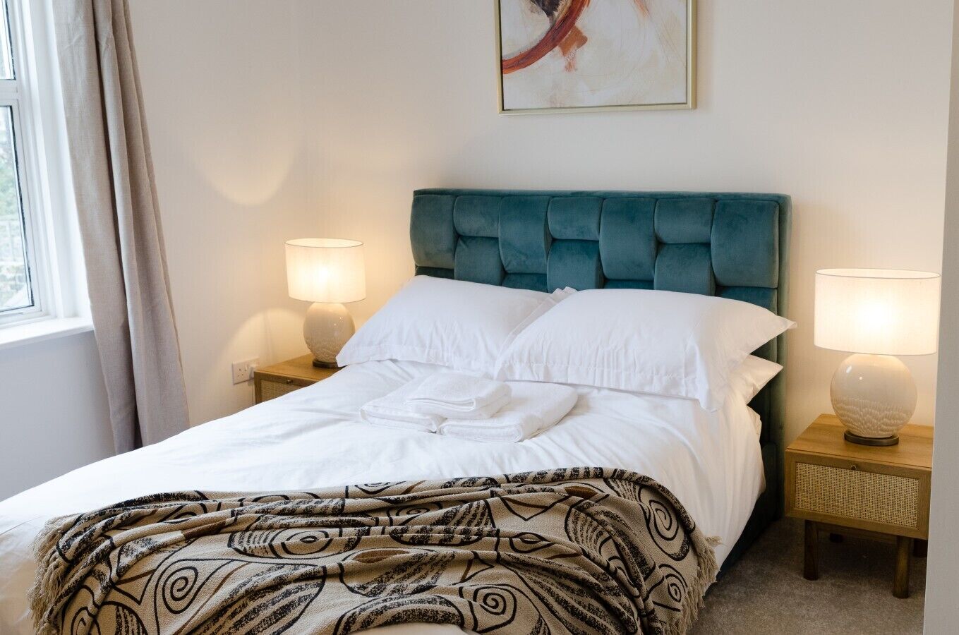 Una cama bien tendida con sábanas blancas, un plaid estampado y un cabecero tapizado en color aguamarina. Hay dos lámparas de noche sobre mesitas de madera.