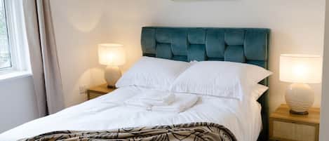 Una cama bien tendida con sábanas blancas, un plaid estampado y un cabecero tapizado en color aguamarina. Hay dos lámparas de noche sobre mesitas de madera.