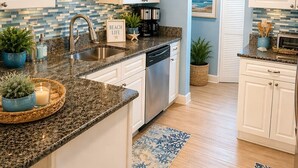 A kitchen with white cabinets, a granite countertop, a coffee maker, and a sign that says 'BEACH LIFE'. - Gorgeous. Direct Beach Access  Heated Oceanfront Pool, Pets OK, No Drive Beach (Daytona Beach Shores)
