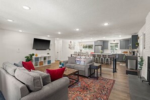 A modern living room with a grey sofa, red cushions, and a coffee table. A kitchen with grey cabinets and a bar area is visible in the background.
