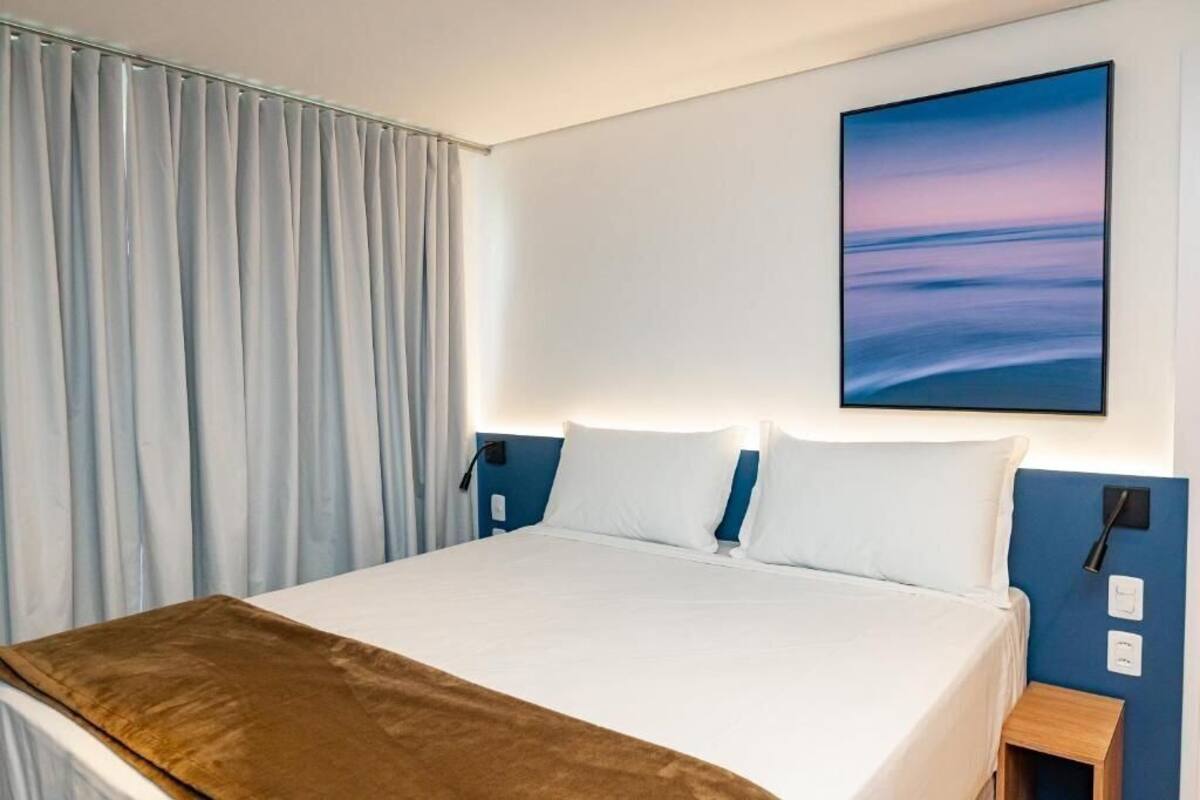 Habitación de hotel con cama, mesita de noche, lámpara de pared y un cuadro de un paisaje.