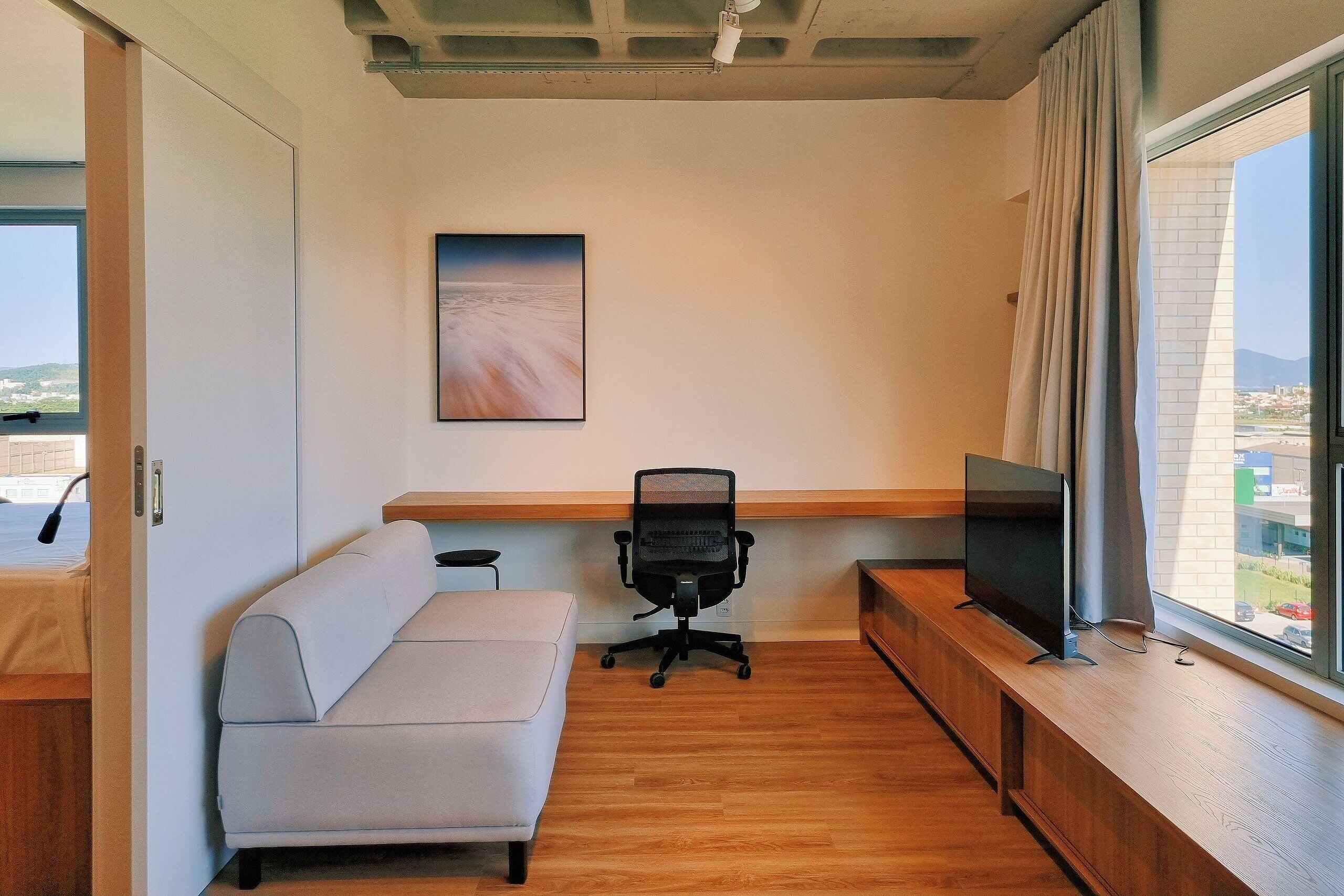A modern office space with a sofa, a desk, a chair, a television, and a large window with a view.
