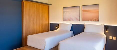 A hotel room with two single beds, a wooden wardrobe, and two framed pictures on the wall.