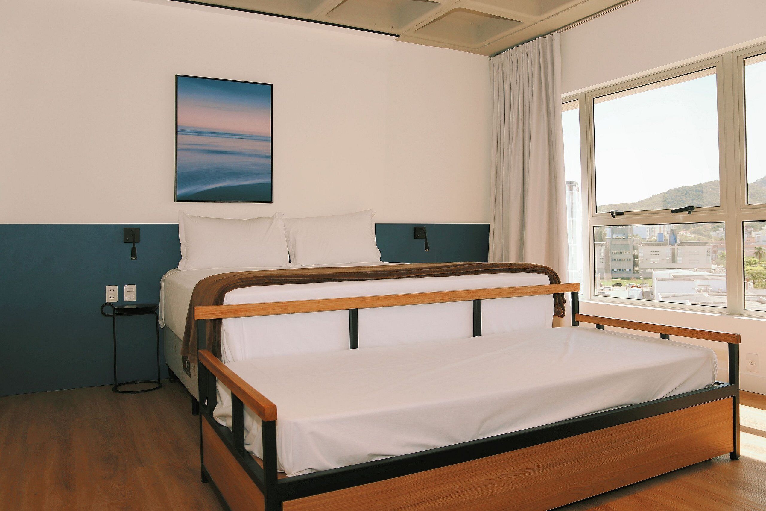 A single bed with a wooden frame and white bedding, a nightstand with a lamp, and a window with a view of buildings and hills.