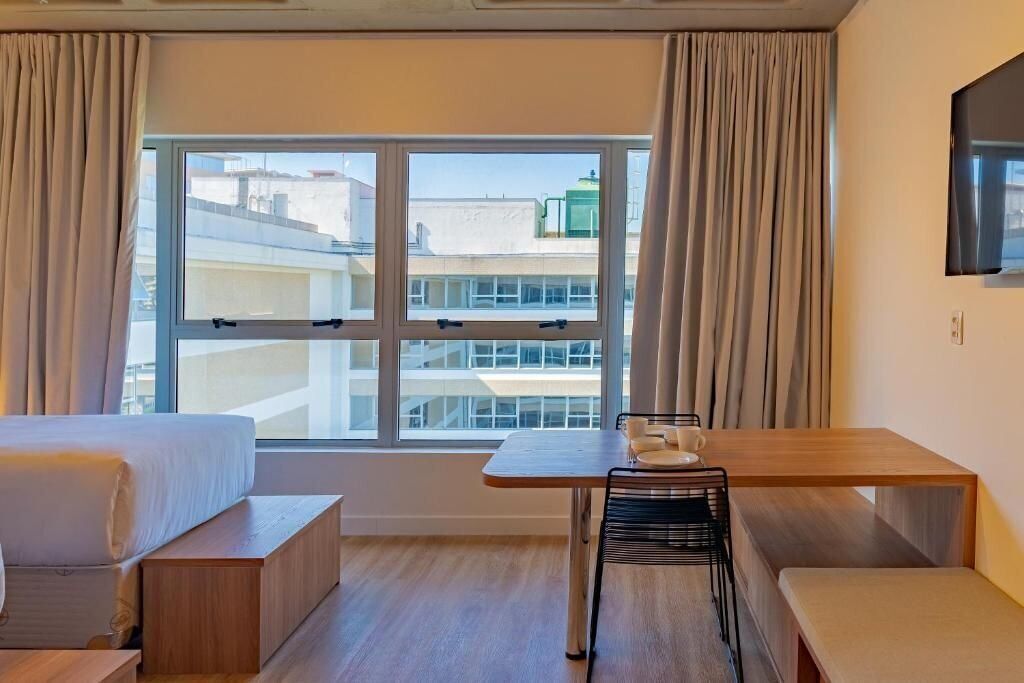 A hotel room with a bed, a small table with a chair, a bench, and a window with curtains.