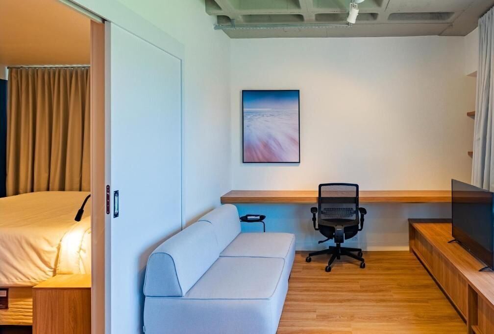 A compact hotel room with a bed, a sofa, a desk with a chair, a television, and a painting on the wall.