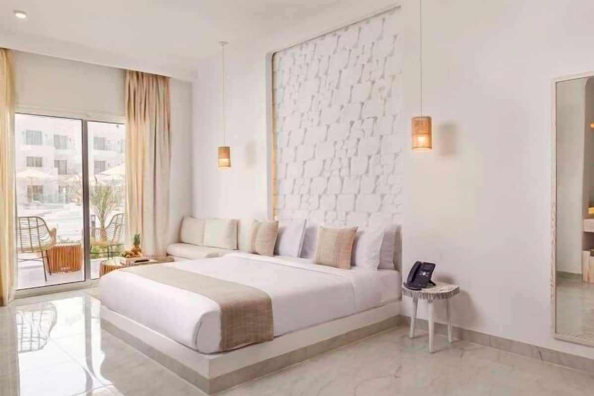 Una habitación de hotel moderna con una cama grande, una mesita de noche pequeña con un teléfono, una lámpara de pared y un ventanal con cortinas.