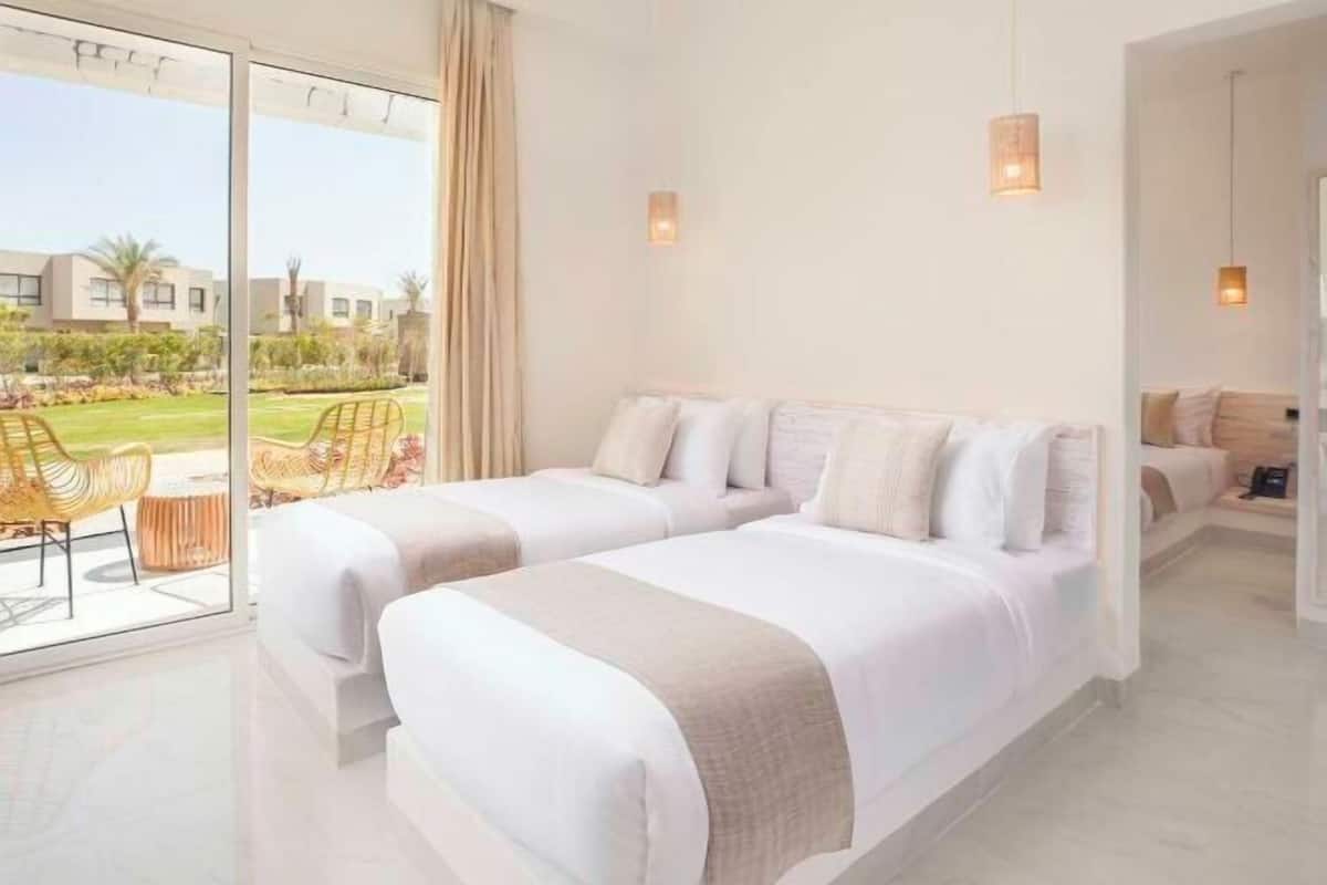 Una habitación de hotel moderna con dos camas, un balcón con mobiliario exterior y vistas de los edificios circundantes.