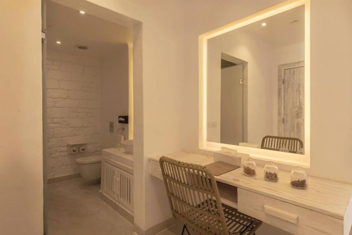 Une salle de bain avec un mur en briques blanches, des toilettes, un meuble-lavabo avec un miroir et une chaise en osier.