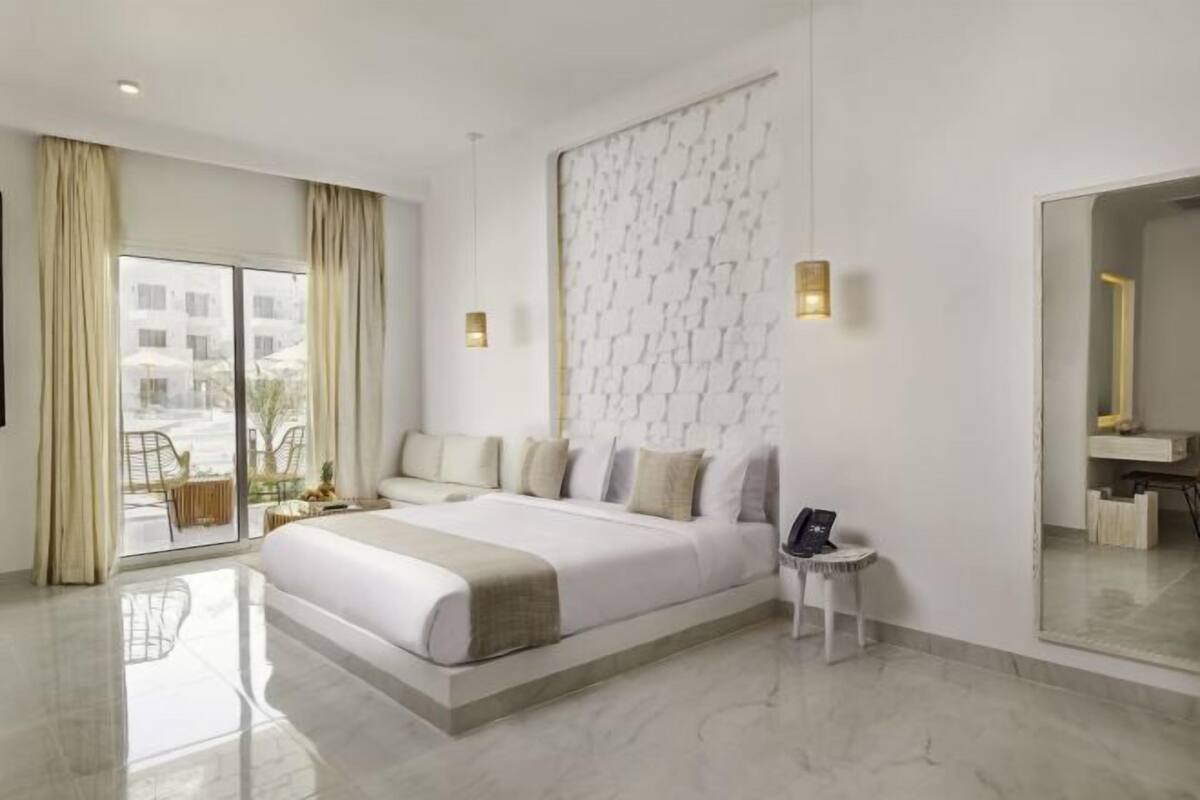 Una habitación de hotel moderna con una cama grande, una mesita auxiliar pequeña, un espejo y vistas al exterior.