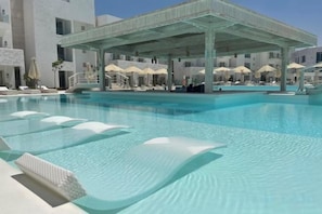 Une piscine avec des chaises longues et des parasols, un espace de repos couvert et un bâtiment en arrière-plan.