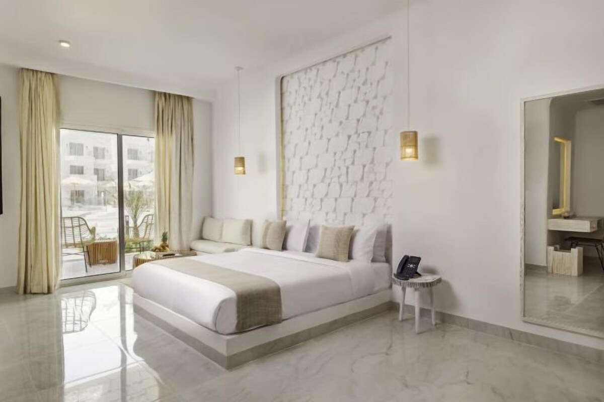Una habitación de hotel moderna con una cama grande, una mesita auxiliar pequeña, un espejo y vistas al exterior.