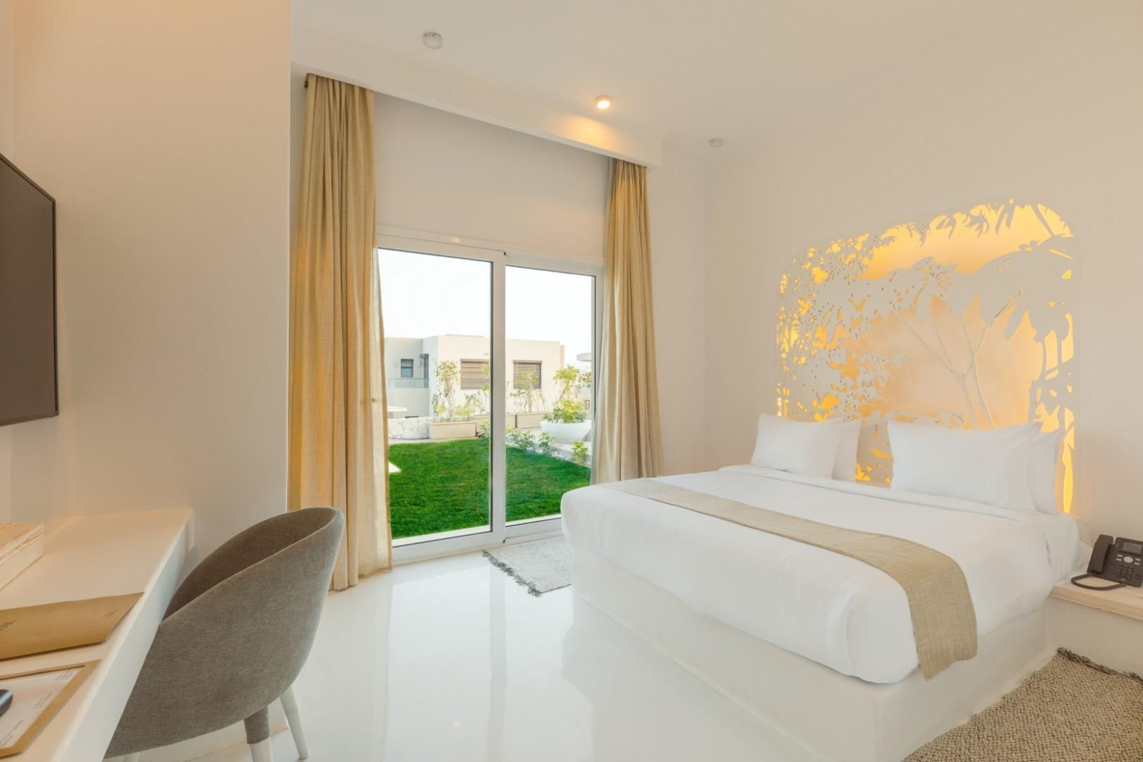 A hotel room with a bed, a desk, a chair, a telephone, and a view of a green lawn and buildings outside.