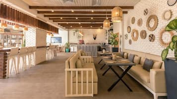 A spacious lounge area with a long wooden bench, a sofa, and a dining table. There is a kitchen area in the background with a bar and various decorative items on the wall.