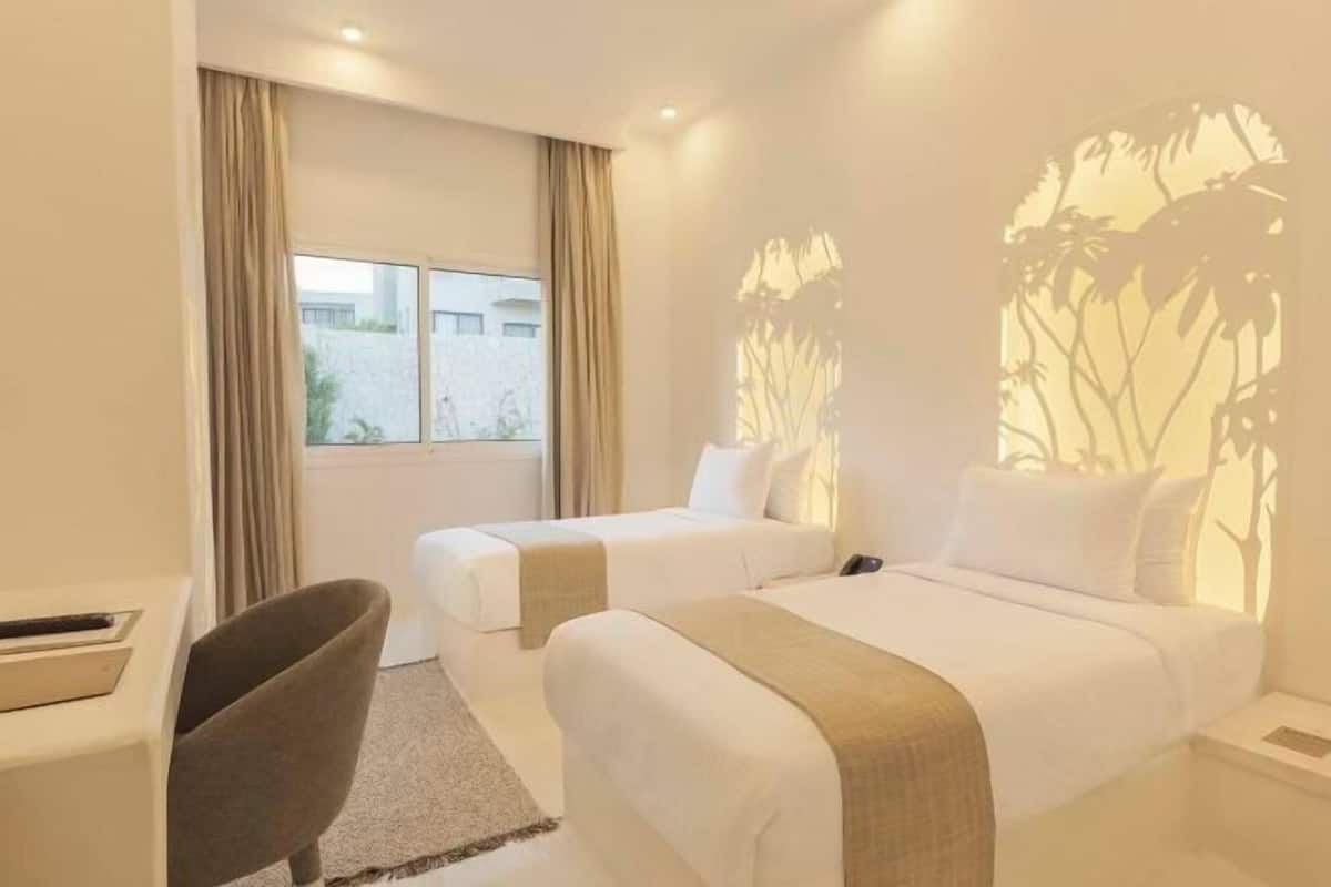 Habitación de hotel con dos camas, un escritorio, una silla y un ventanal con cortinas.