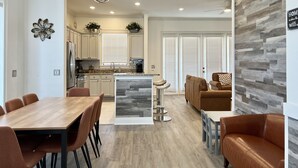 A modern kitchen with white cabinets, a central island, and pendant lighting. There is a dining area with a wooden table and chairs, and a living room with a sofa and a coffee table.