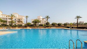 A swimming pool with clear blue water, surrounded by a fence, and adjacent to a row of white buildings with balconies and palm trees. - Stunning Ground Floor Apartment (Sucina)