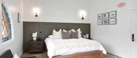 Un dormitorio con cama, mesitas de noche, una silla, mesita auxiliar, luz de pared e imágenes enmarcadas.