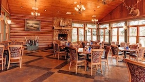 A rustic dining area with wooden walls, a stone fireplace, and large windows. Tables and chairs are arranged for dining. - Balsam Lake Lodge (Balsam Lake)