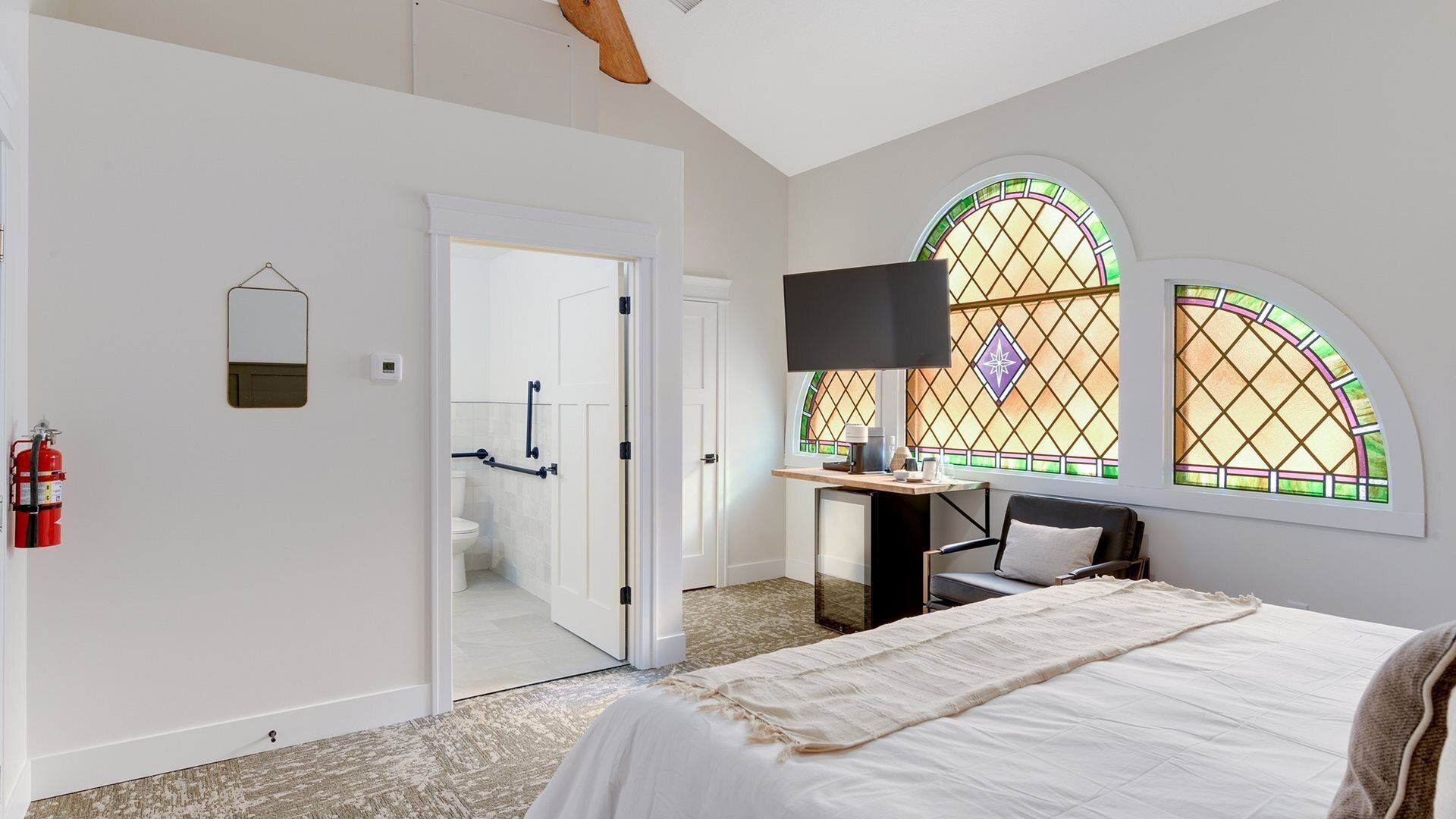 Un dormitorio con cama, escritorio con silla, televisión y dos ventanas con vitrales.