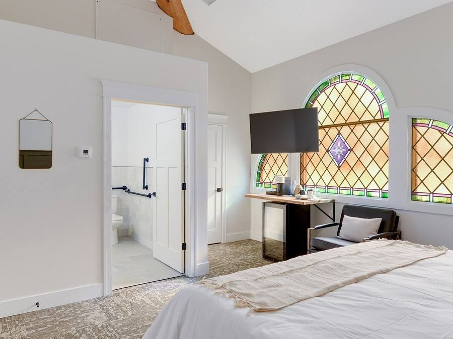 Un dormitorio con cama, escritorio con silla, televisión y dos ventanas con vitrales.