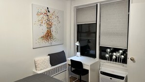 Een compacte, moderne slaapkamer met een wit bureau, een zwarte stoel, een witte radiator en een groot raam met zonwering.