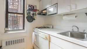 Een kleine keuken met witte kasten, een gasfornuis, een roestvrijstalen spoelbak en een raam met uitzicht op een bakstenen gebouw.