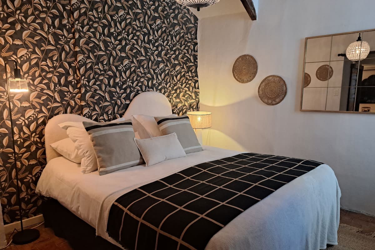 Una cama impecablemente hecha con colcha a cuadros blanco y negro, cabecera blanca y múltiples almohadas. Se observa un espejo con marco decorativo fijado a la pared y una lámpara colgante.
