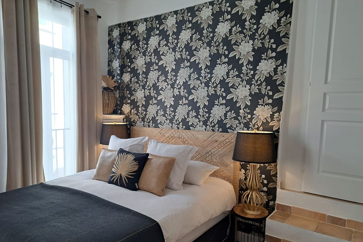Una cama bien hecha con un edredón oscuro y varias almohadas, una pared con estampado floral, una lámpara en una mesita de noche y una ventana con cortinas.