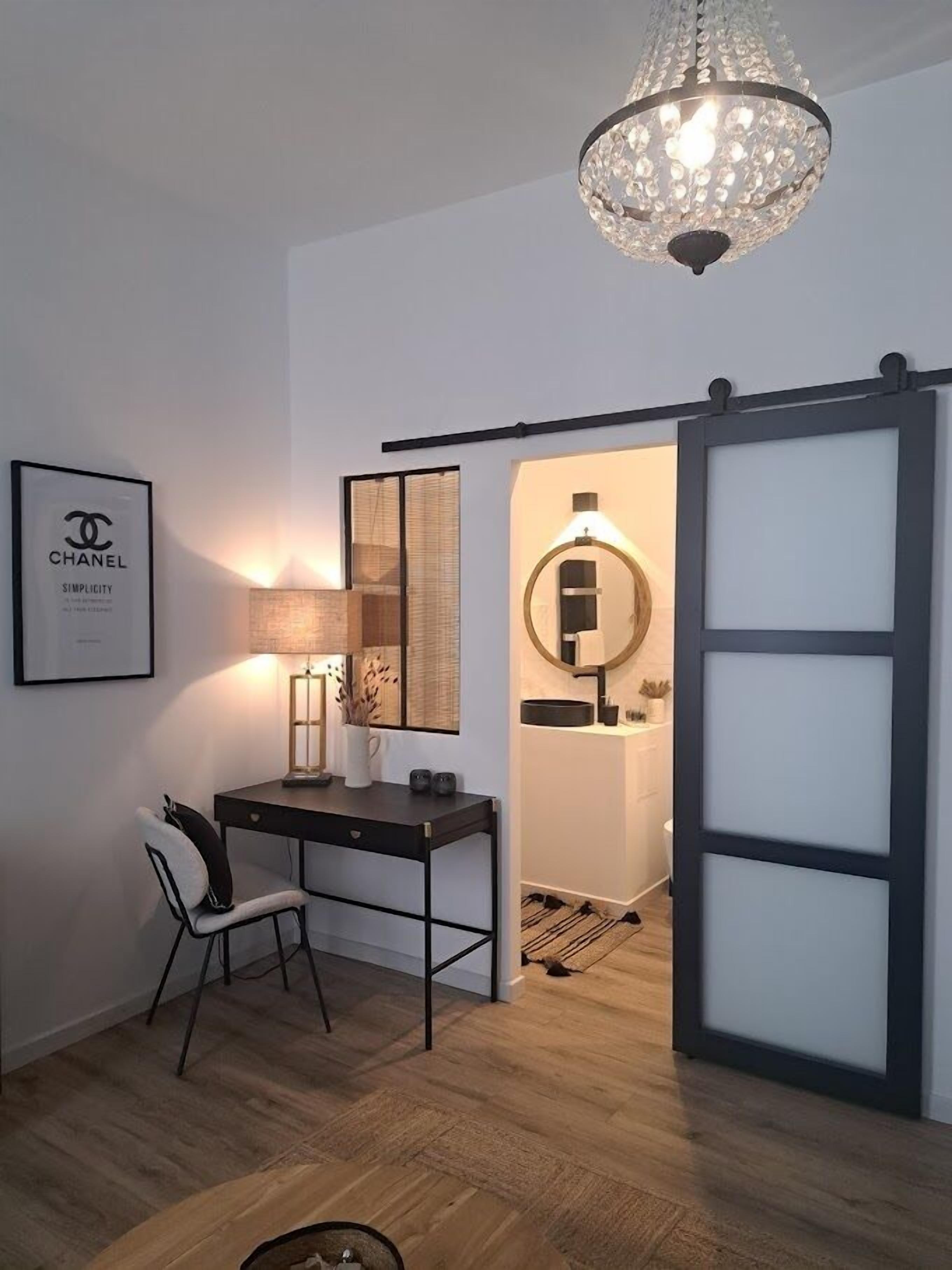 Una habitación moderna con escritorio, silla y candelabro. Hay un cartel de Chanel enmarcado en la pared y un espejo con marco redondo.