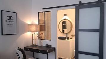 Una habitación moderna con escritorio, silla y candelabro. Hay un cartel de Chanel enmarcado en la pared y un espejo con marco redondo.