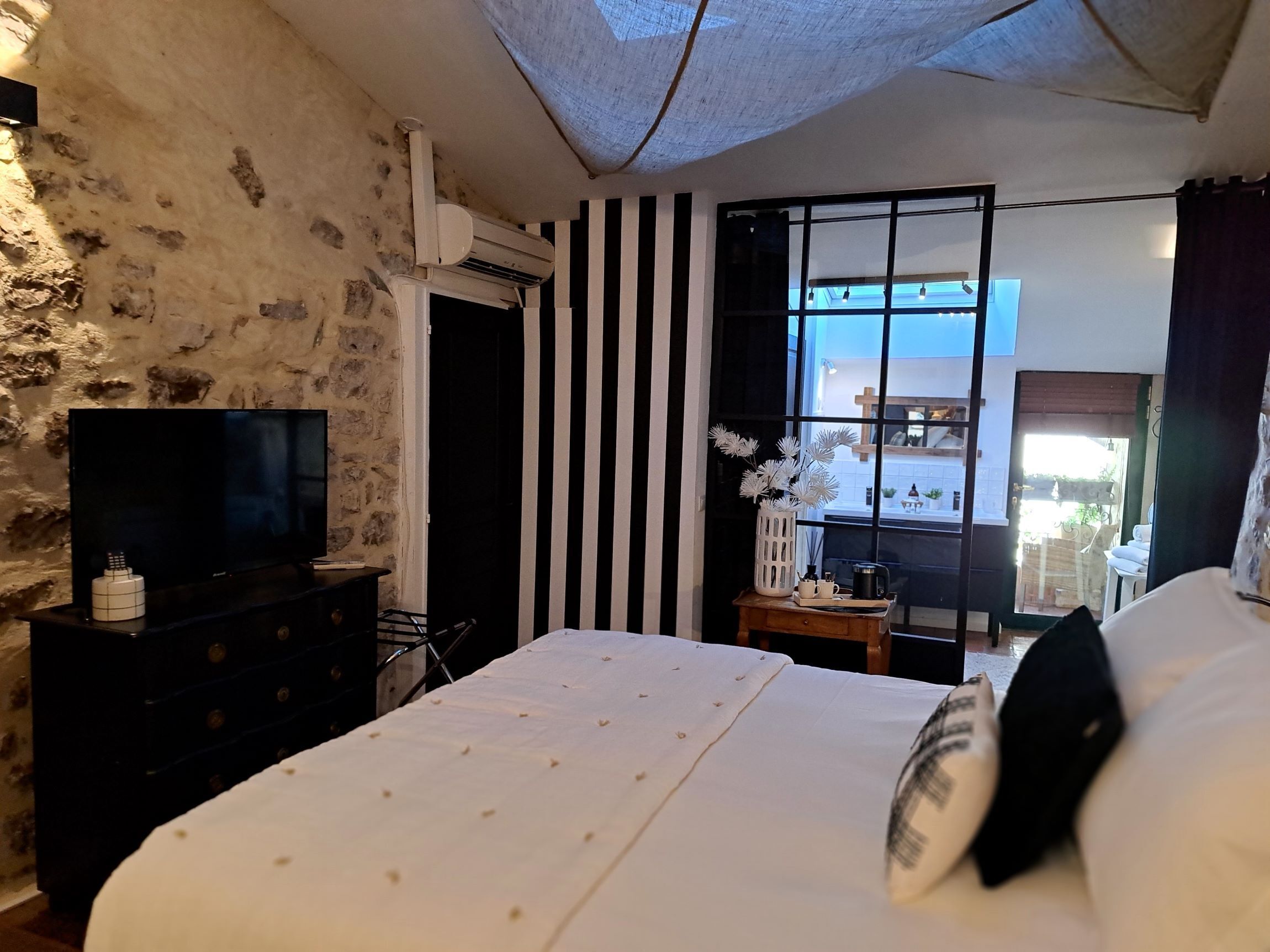 Ein Schlafzimmer mit Steinwand, einem Flachbildfernseher auf einer Kommode, einem Bett mit weißer Bettwäsche, einem Fenster mit schwarz-weißen Streifen und Blick nach draußen.