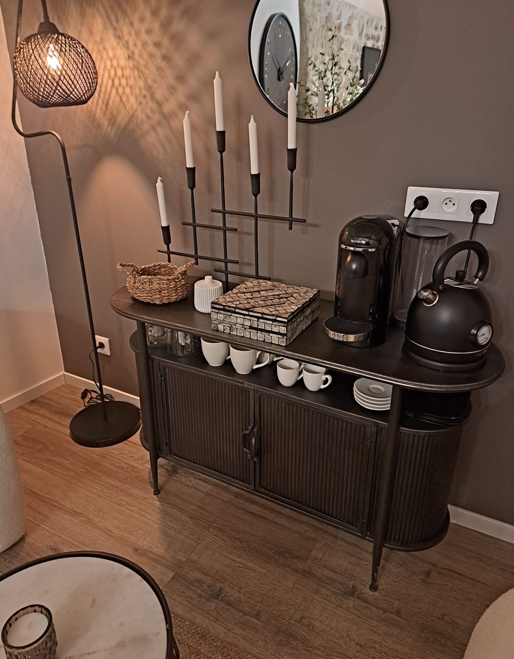 A dark wooden console table with a mirror, candles, a basket, a coffee maker, a kettle, and several cups.