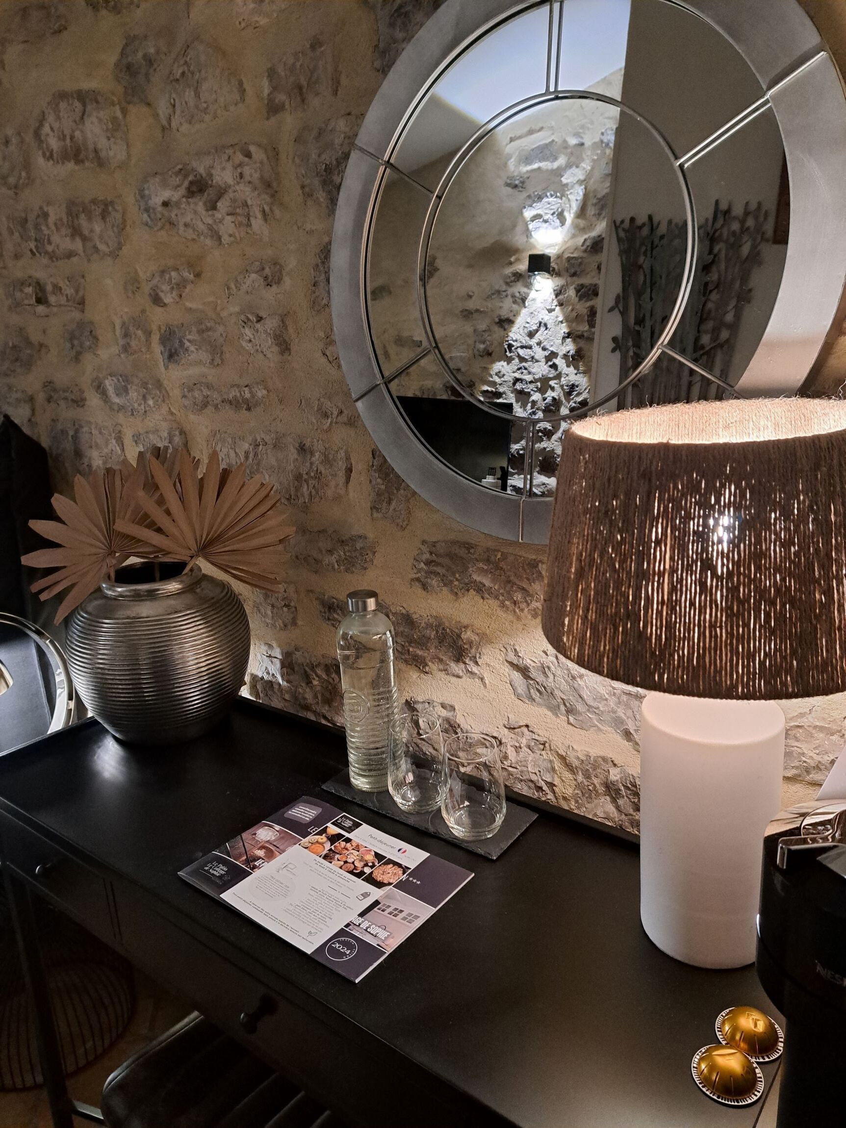 A modern interior with a round mirror, a textured lamp, a vase, and a menu on a dark countertop against a stone wall.