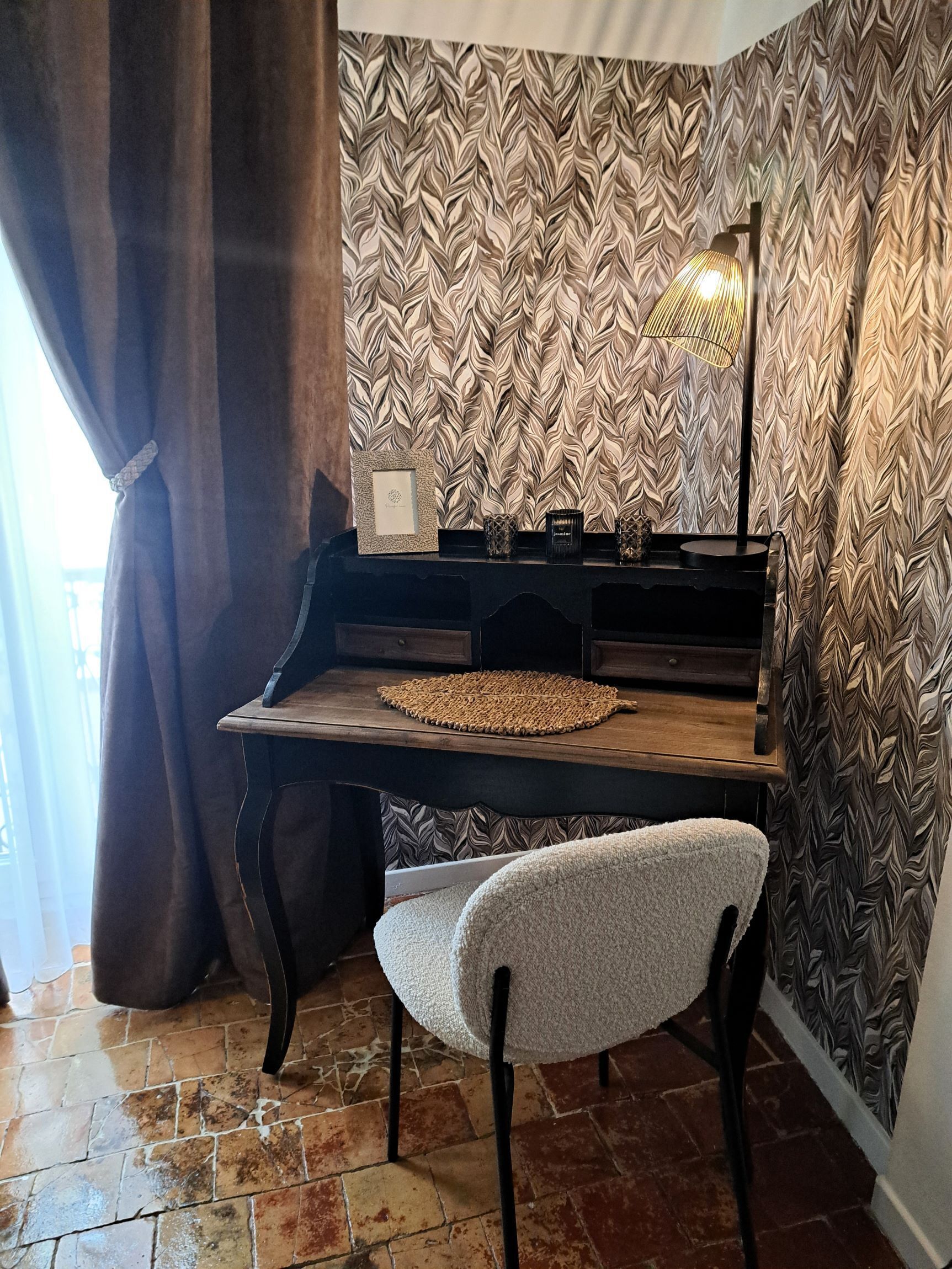 A room with a wooden desk, a chair, a wall with a leaf-patterned wallpaper, a floor lamp, and a curtain.