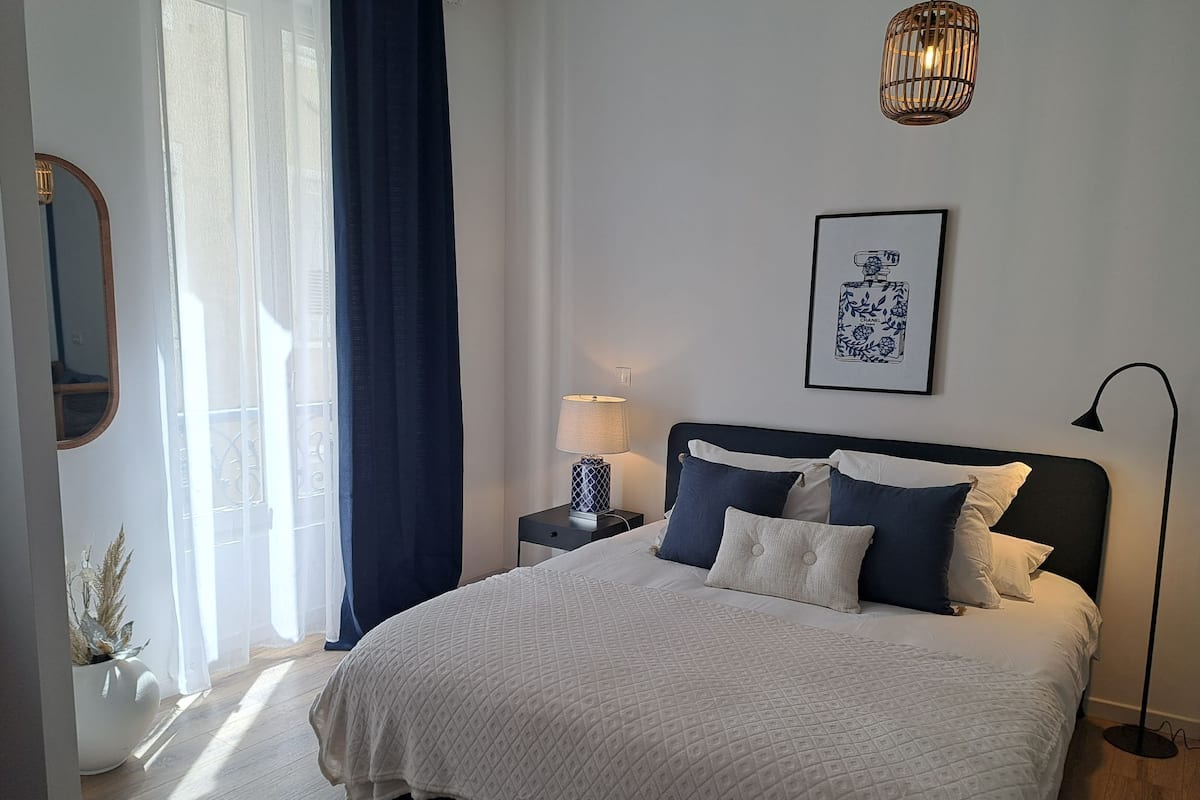 Un dormitorio ordenado con una cama grande, una mesita de noche con lámpara, un cuadro en la pared y una ventana con cortinas.