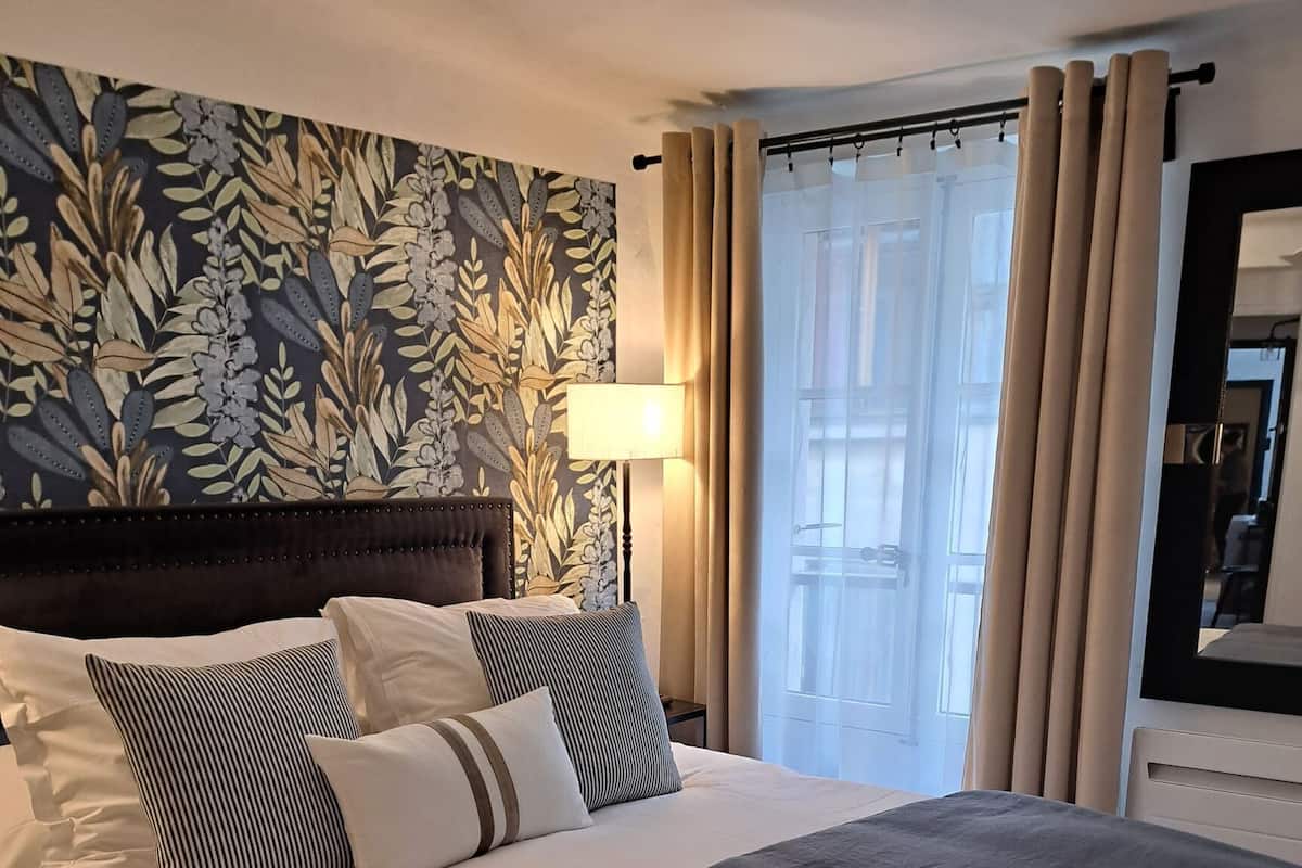 Una cama bien hecha con edredón gris y almohadas a rayas, papel tapiz floral, una lámpara de noche y vistas al exterior a través de una ventana con cortinas.
