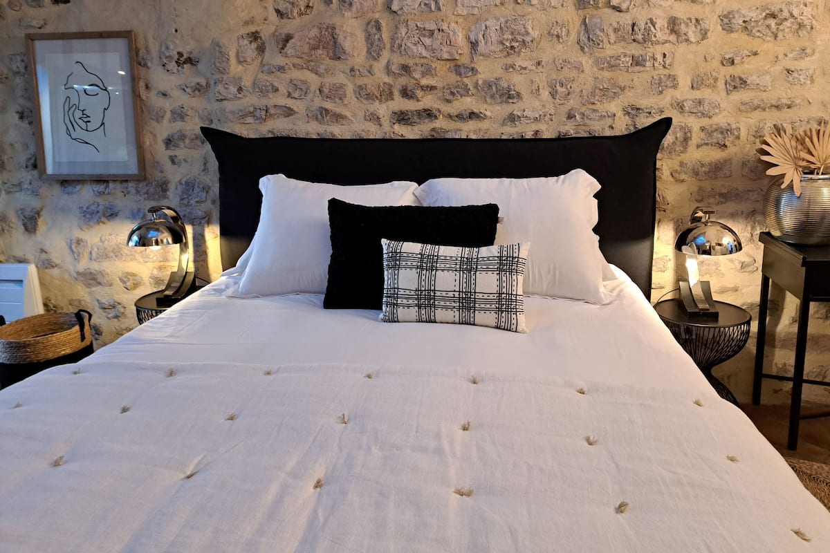 Un dormitorio con pared de piedra, cabecero negro, ropa de cama blanca y dos mesitas de noche con lámparas.