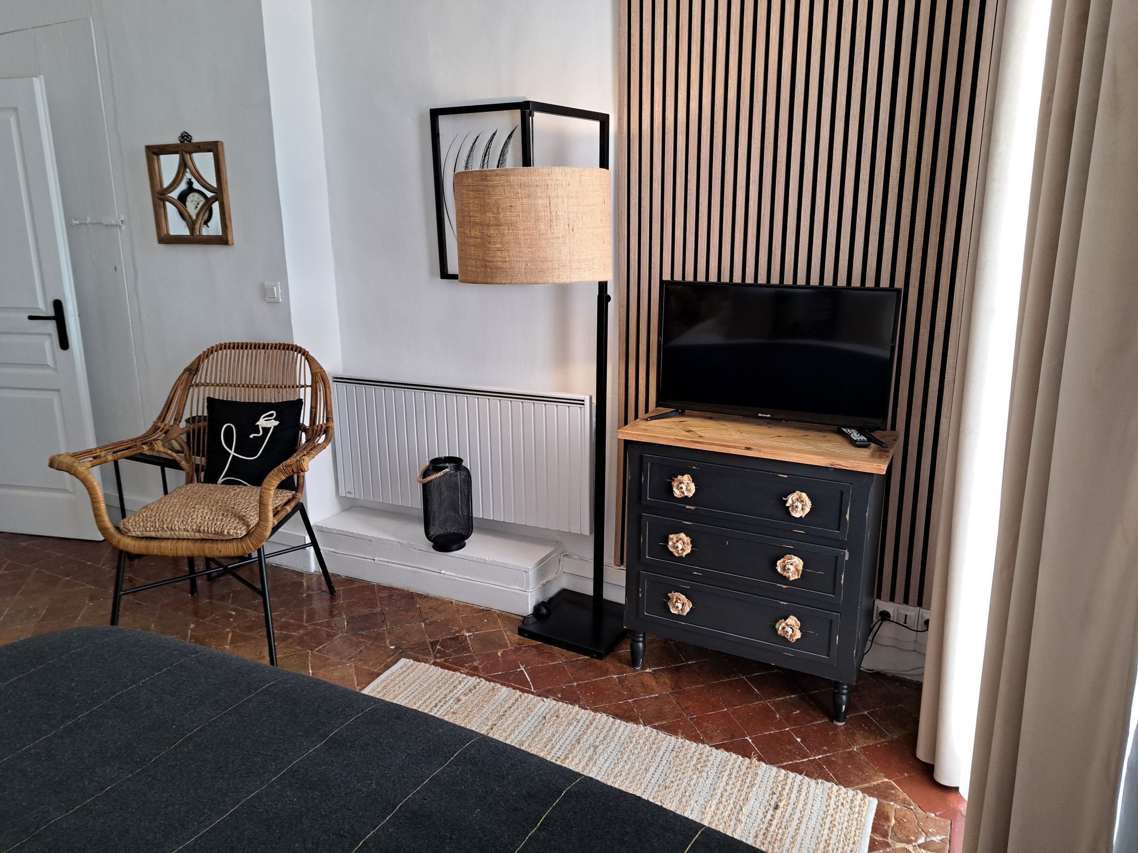 Una habitación con una televisión sobre un mueble de madera, una silla de mimbre, una lámpara negra y un radiador.