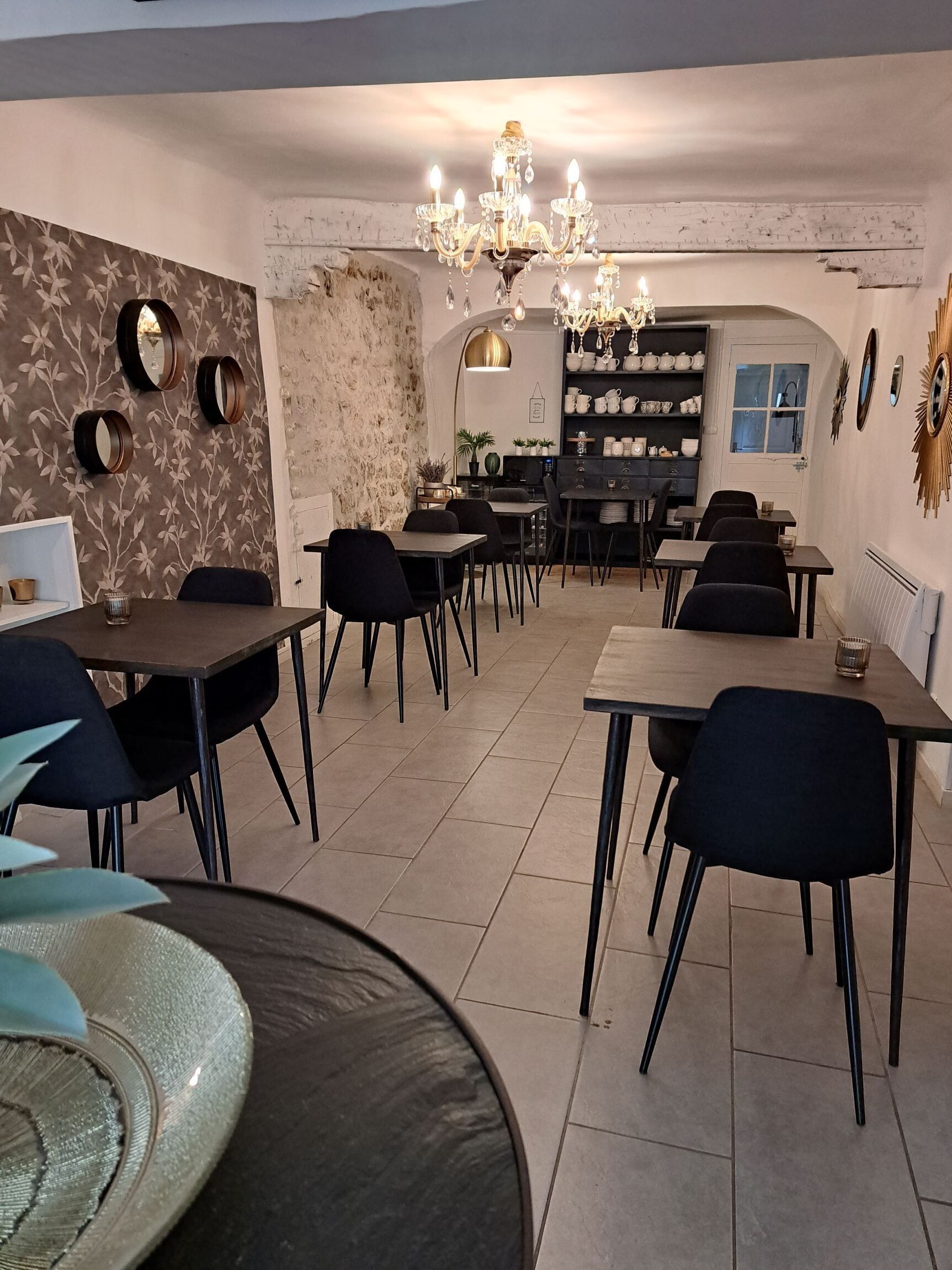 A dining area with round tables, black chairs, and a chandelier. There are wall-mounted mirrors and a shelf with teapots.