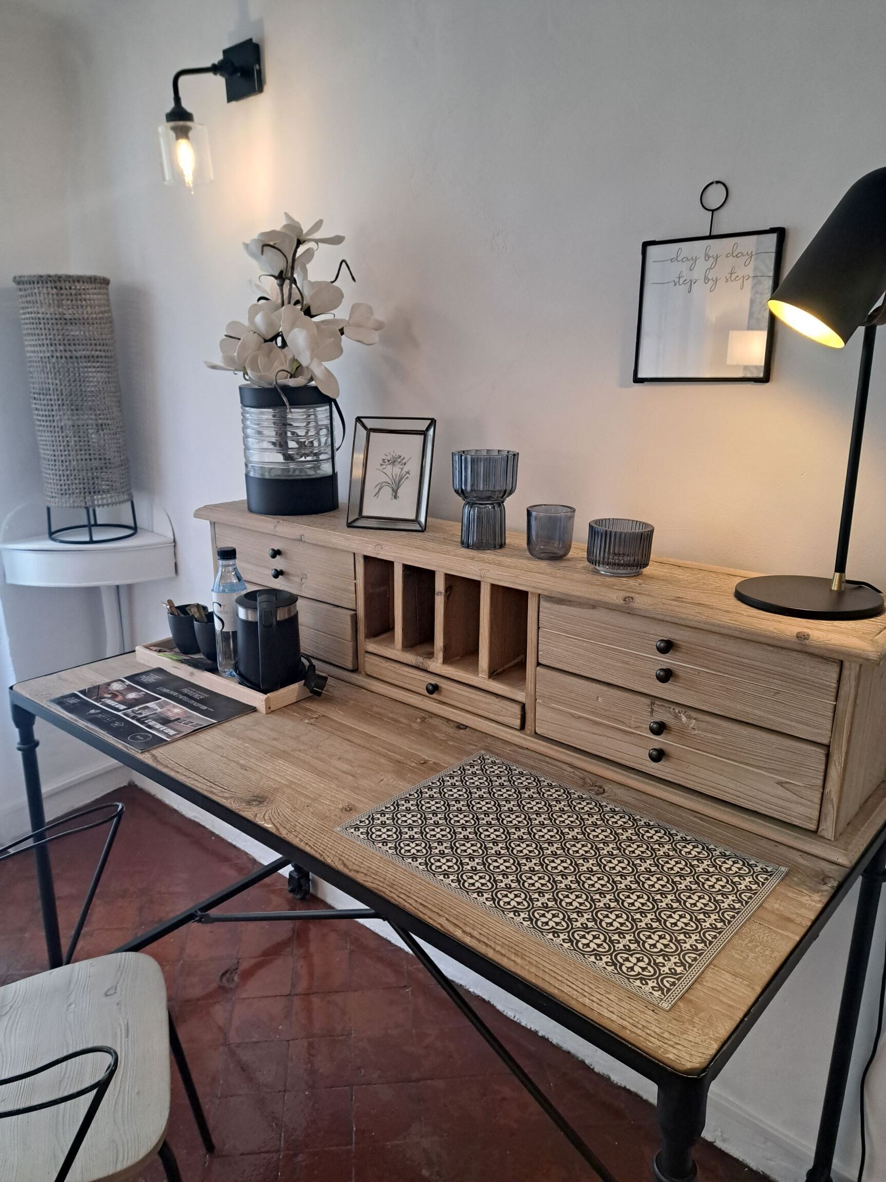 A wooden desk with drawers, a framed picture, a vase with white flowers, a black lamp, and a small table with a lamp.