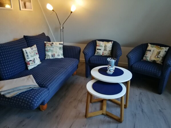 A living room with a blue sofa set, two armchairs, a small white table with blue cushions, and a floor lamp. - Scharhörn House, Vacation Apartment 14 (Cuxhaven)
