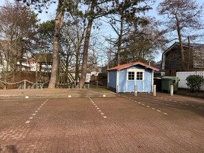 Une aire de stationnement dallée avec un petit bâtiment bleu, qui pourrait être un abri utilitaire, et une zone clôturée avec des arbres.